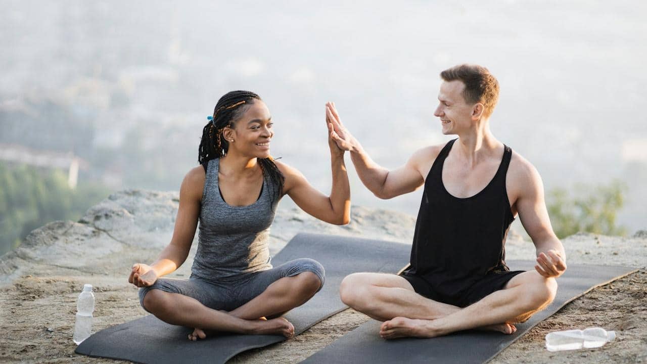 A couple high-fiving each other while sitting cross-legged on yoga mats on top of a cliff.