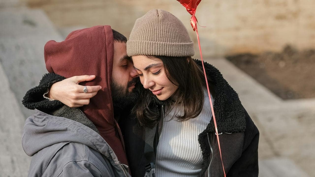 A couple cuddles closely outdoors, sharing a quiet moment while holding a red heart balloon.