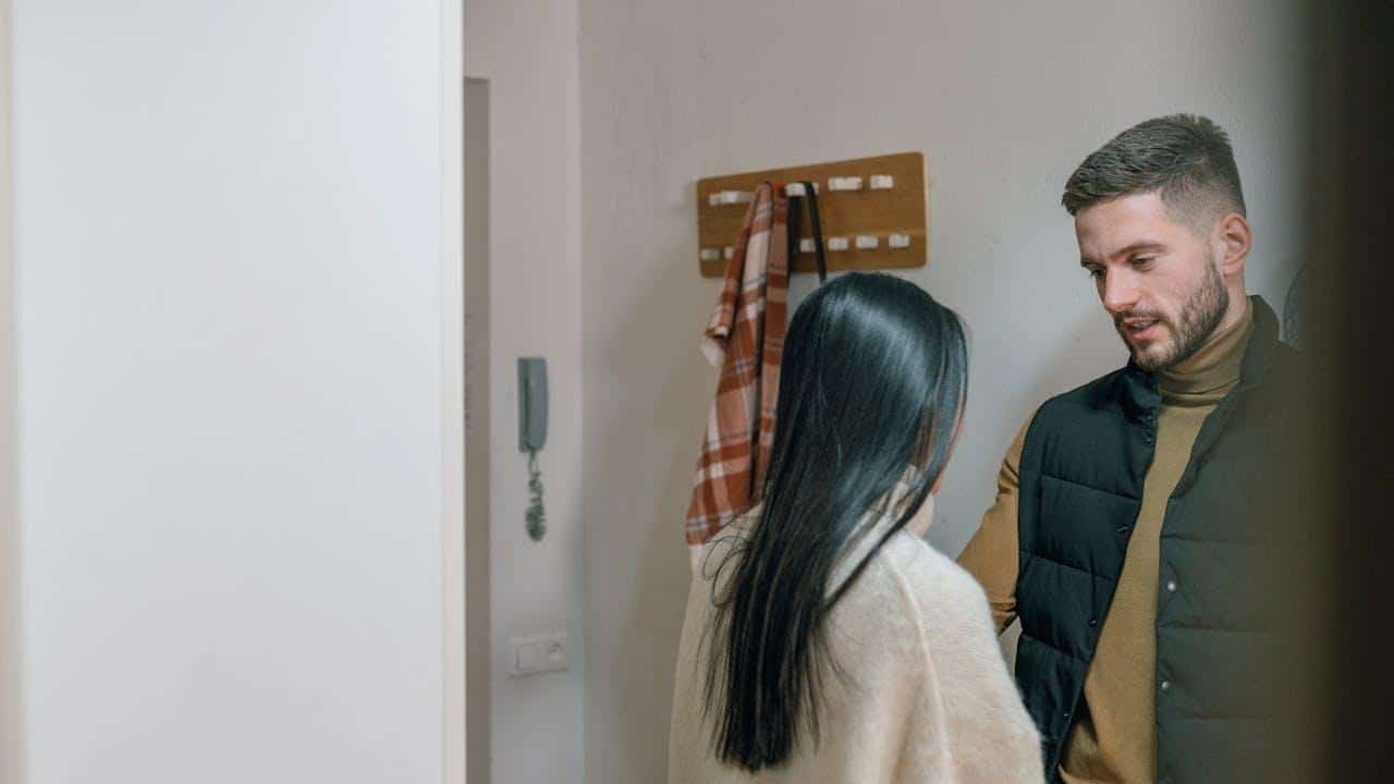 A man with short hair looks toward a woman with long dark hair in a hallway.
