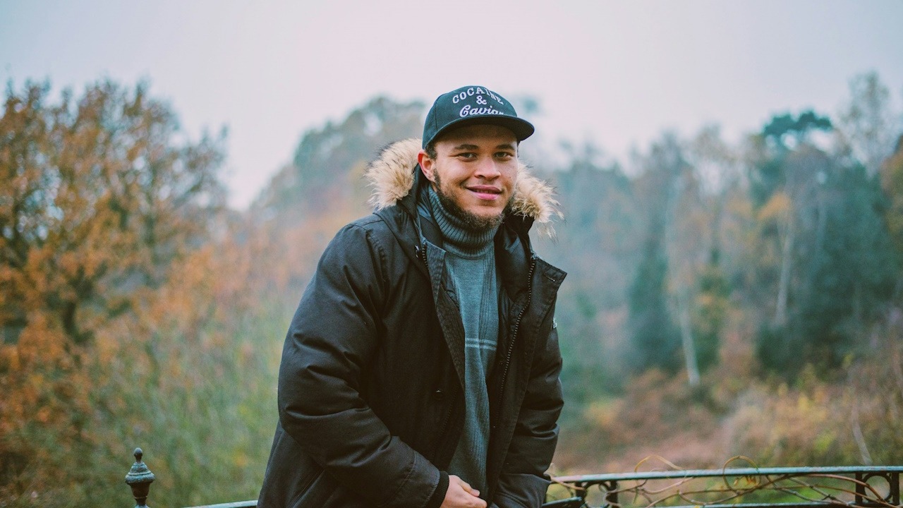 A man wearing a cap and smiling at the camera with a picturesque view behind him.