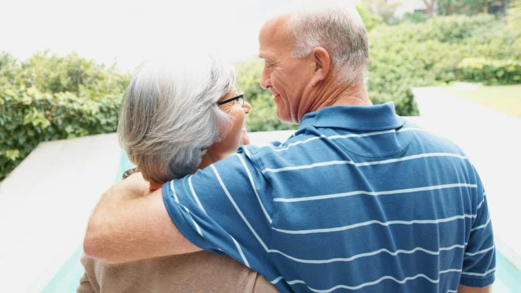 An older couple smiling at each other while standing outdoors with arms around one another.