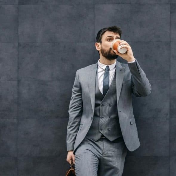 A man in a gray suit sipping coffee against a dark wall.