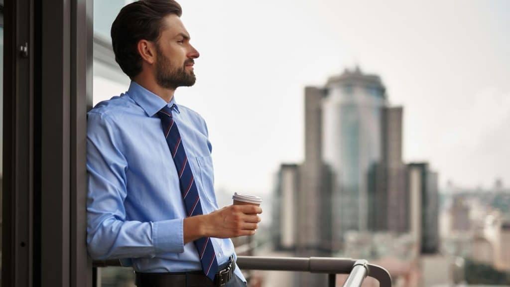 A businessman holding a coffee cup while looking out over a city from a balcony.
