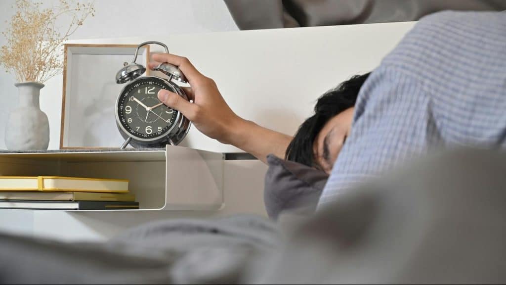 A person reaching to turn off an alarm clock on a bedside table.