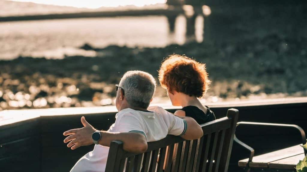 A couple sits on a bench by the water, watching the sunset.
