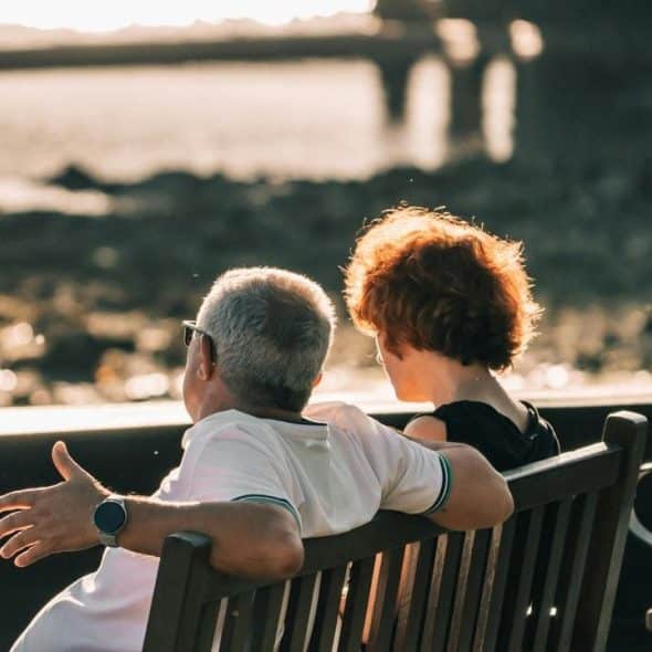 A couple sits on a bench by the water, watching the sunset.
