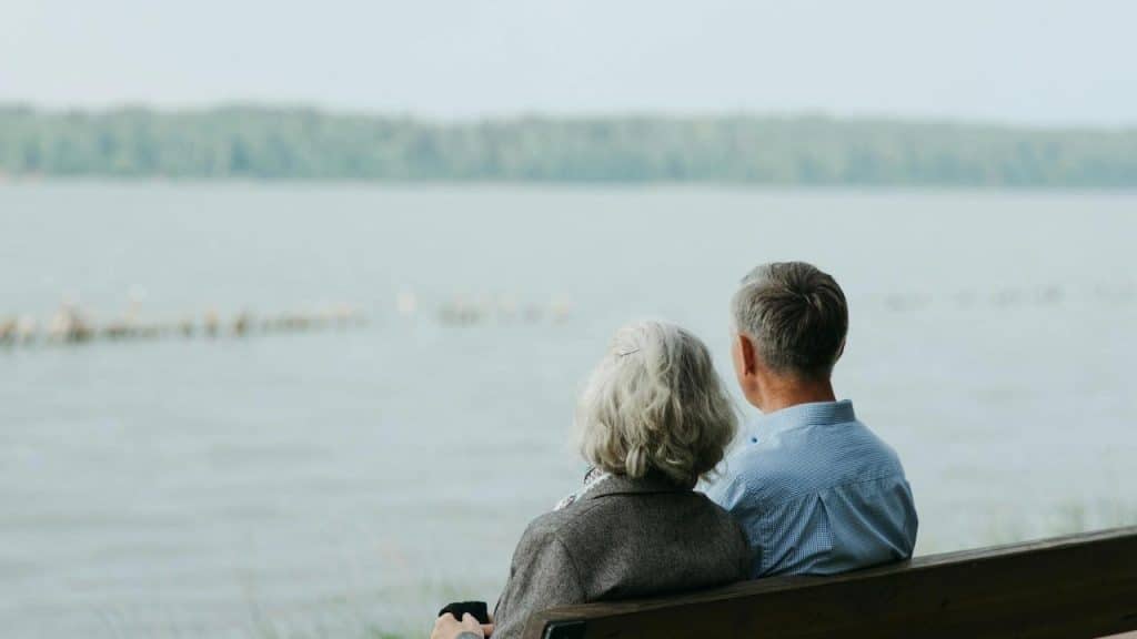 An older couple sitting on a bench and gazing out over a calm lake.