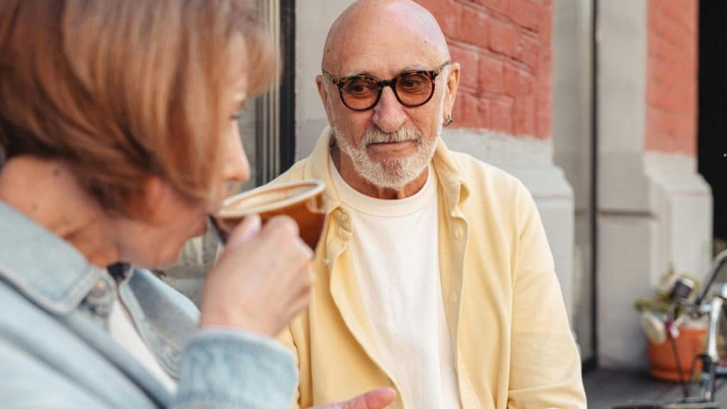 An older man watching a woman drink coffee at an outdoor café.