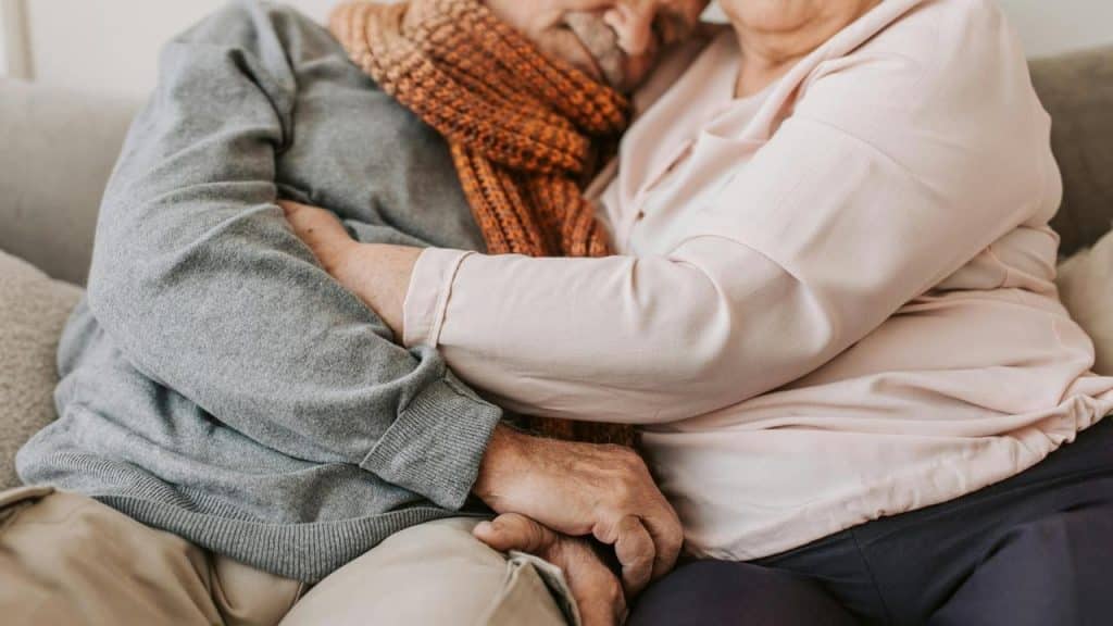 An older couple cuddling closely on a couch with their hands intertwined.