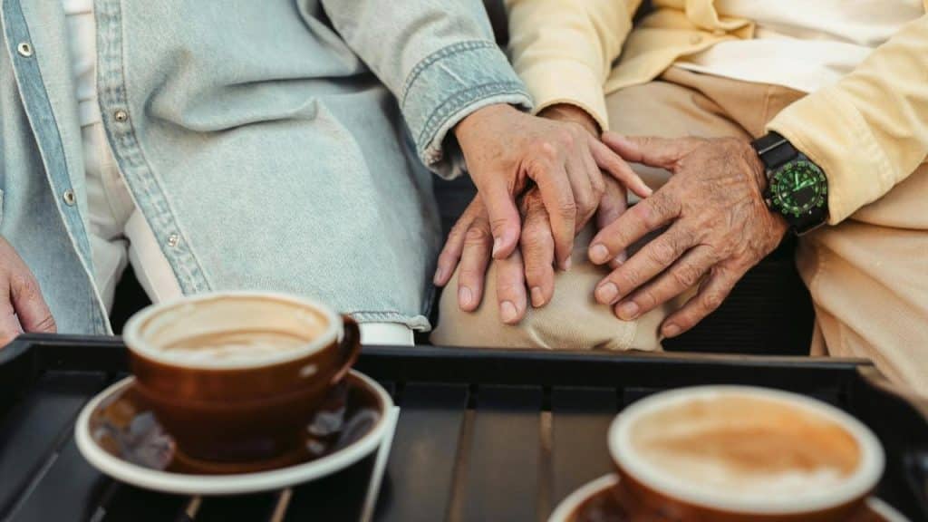 An older couple’s hands resting gently together beside coffee cups.