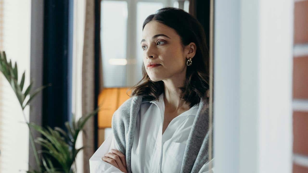 A woman standing indoors and looking thoughtfully out of frame.