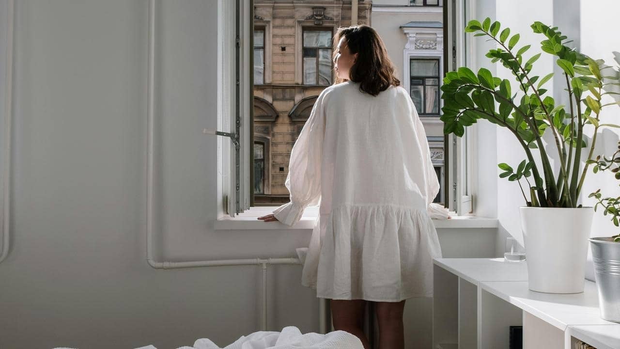 A person in a light dress standing by an open window and looking outside.