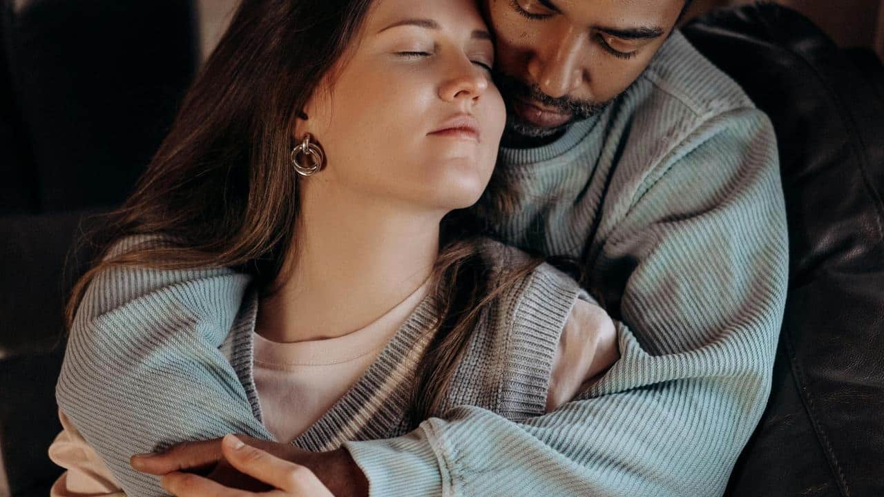 A couple embracing closely on a couch with one person resting their head against the other.
