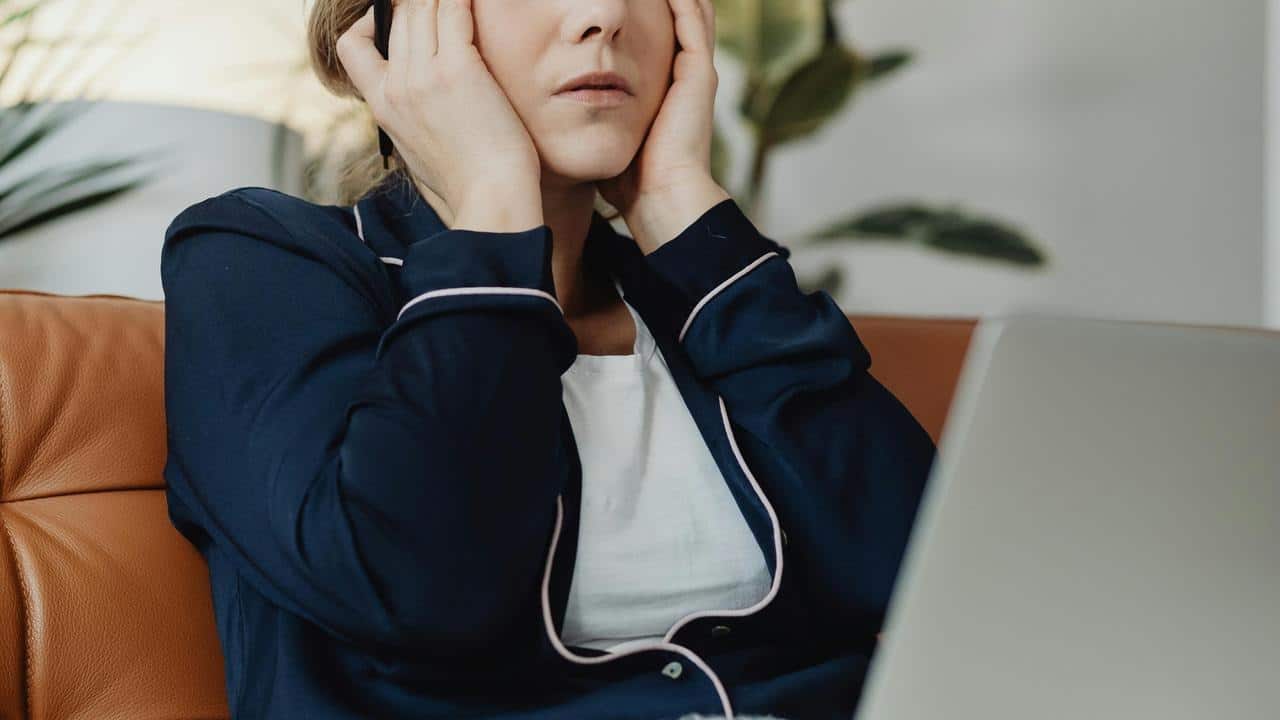 A person holding their head while sitting on a couch with a laptop.