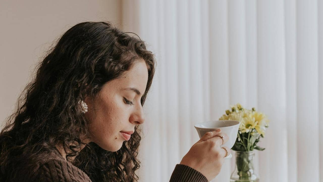 A woman holding a cup and looking down beside a vase of flowers indoors.