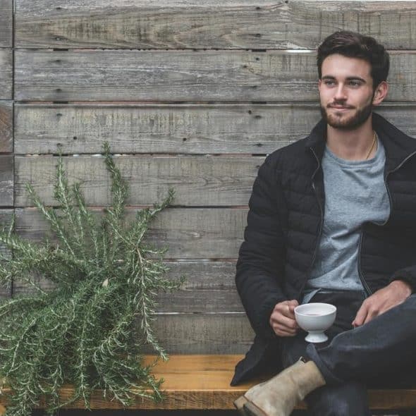 A man sits on a bench holding a cup in front of a wooden wall.