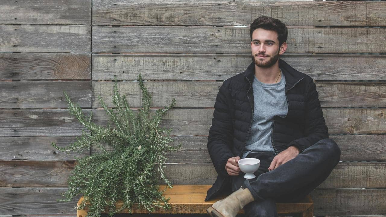 A man sits on a bench holding a cup in front of a wooden wall.