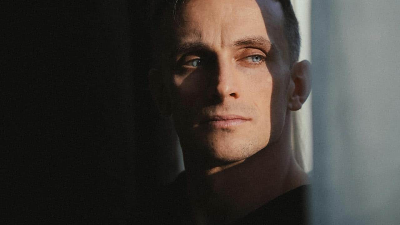 A close-up of a man gazing sideways, with part of his face in shadow near a window.