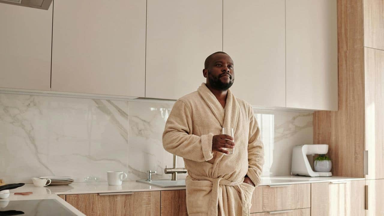 A man in a bathrobe standing in a kitchen holding a glass.