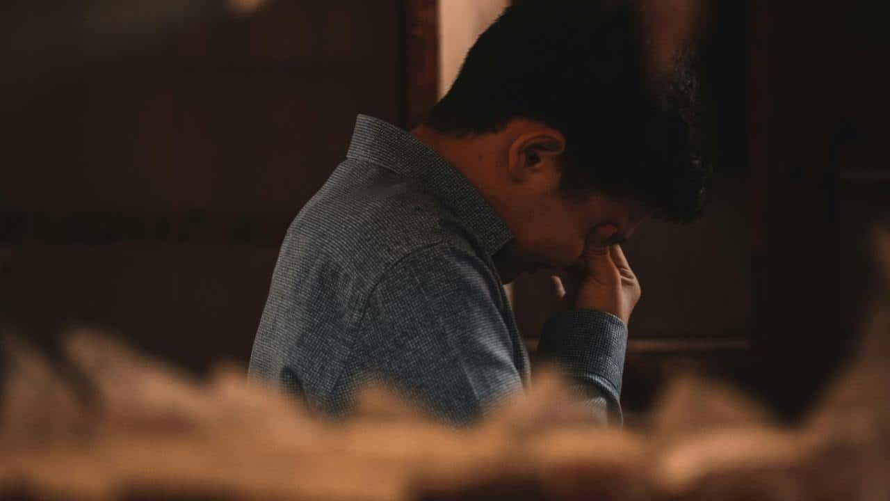 A man sitting indoors with his head bowed and his hand covering his face.