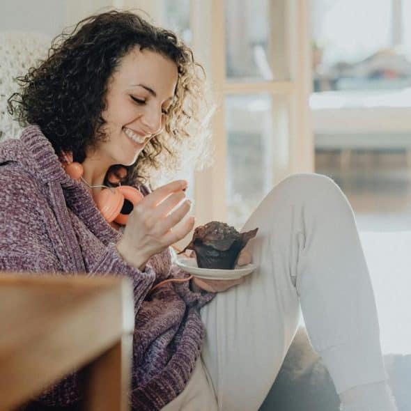 A smiling woman sits indoors eating a muffin with headphones around her neck.