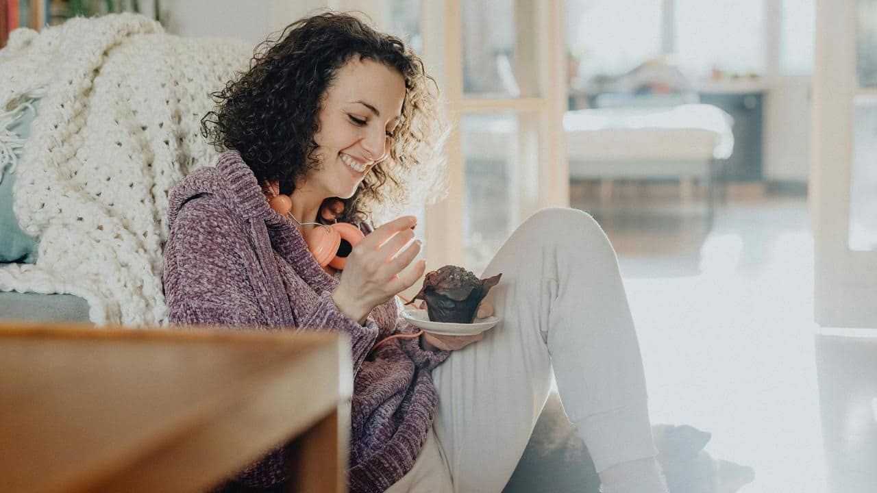 A smiling woman sits indoors eating a muffin with headphones around her neck.