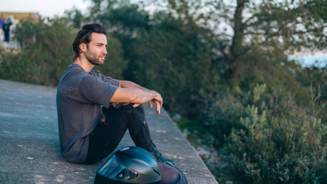A man sits outdoors looking into the distance with a motorcycle helmet nearby.
