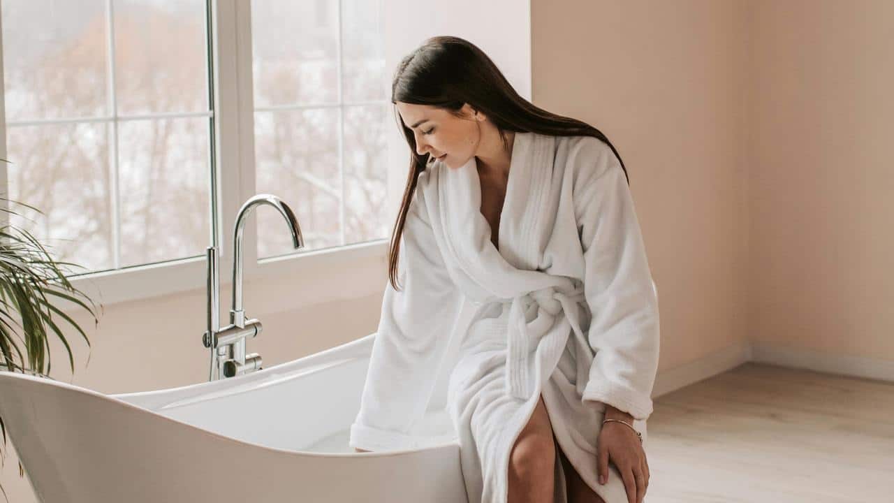 A woman in a white bathrobe tests the water in a bathtub.