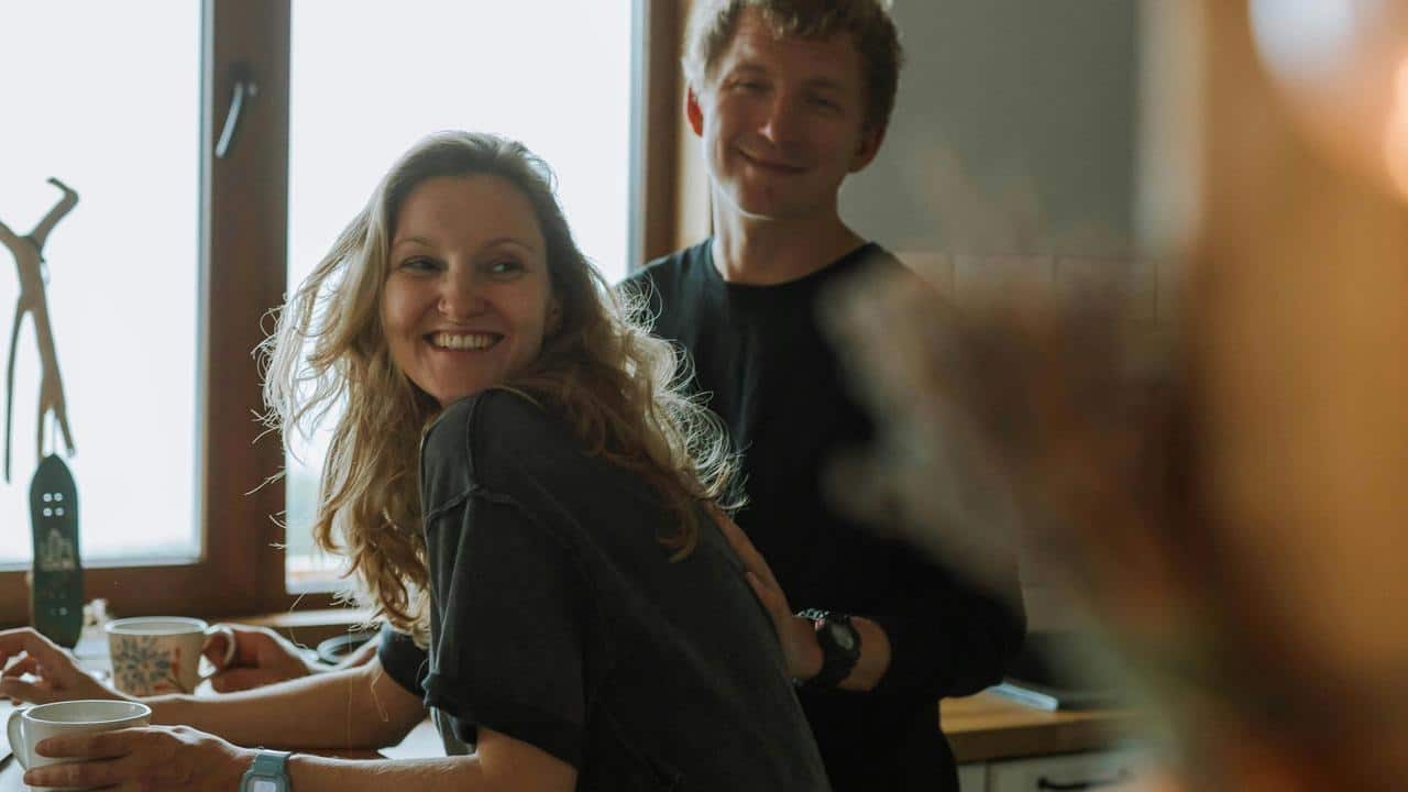 A smiling couple stands together in a cozy kitchen near a window.