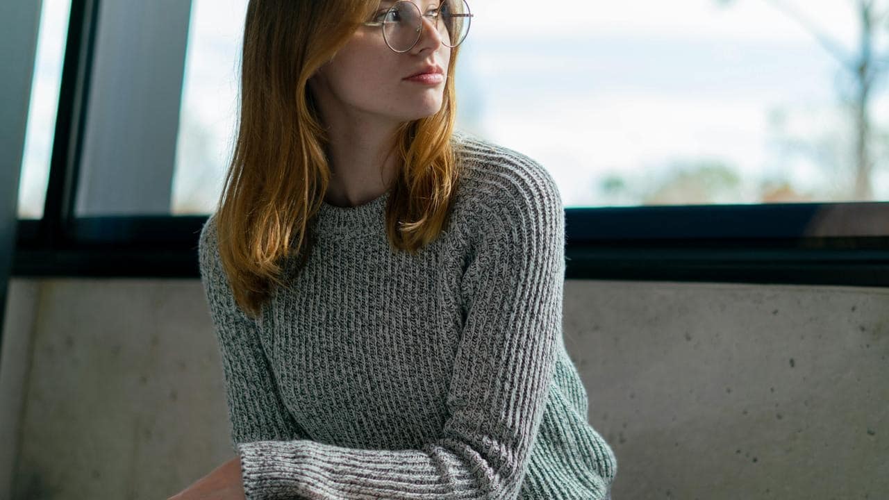 A woman wearing glasses and a gray sweater looks to the side.