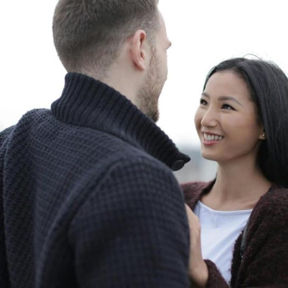 A smiling woman gazes at a man as they stand close together outdoors.