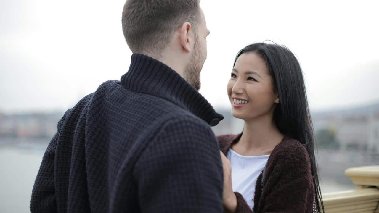 A smiling woman gazes at a man as they stand close together outdoors.