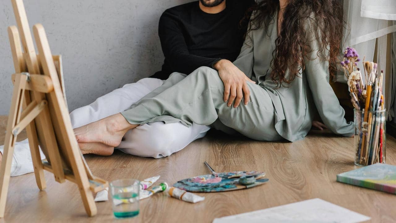 A couple sitting on the floor near an easel, surrounded by art supplies.