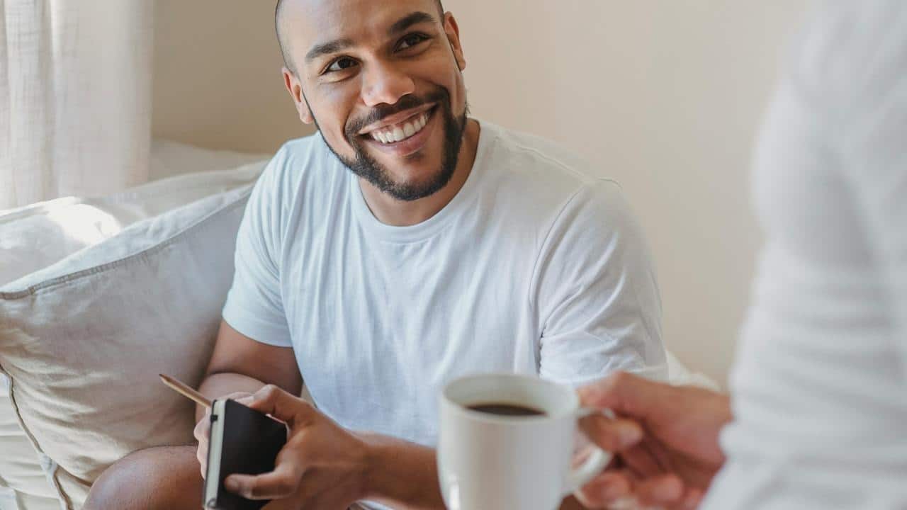 A smiling man sitting on a couch, holding a phone and looking at someone offering coffee.