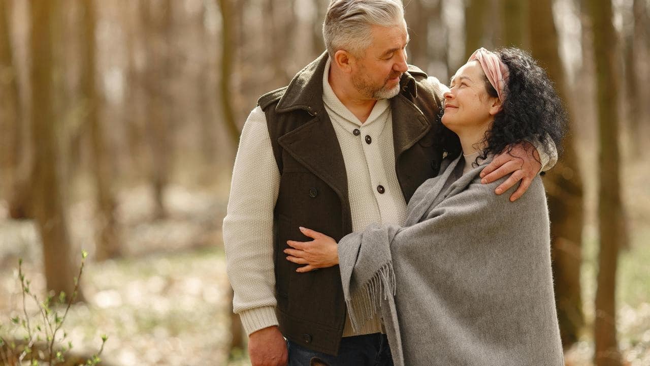 A couple standing close together in a forest, looking at each other affectionately.