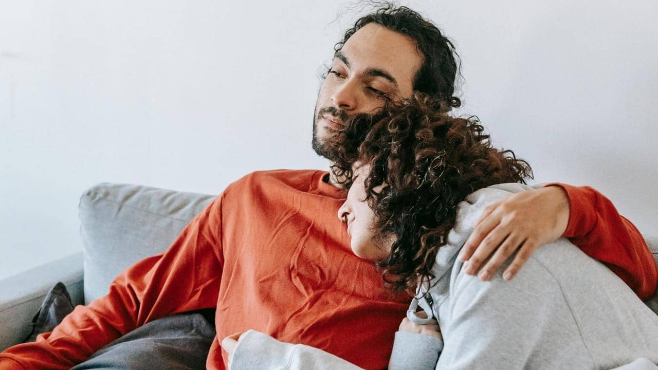 A couple sitting on a couch, with one resting their head on the other’s shoulder.
