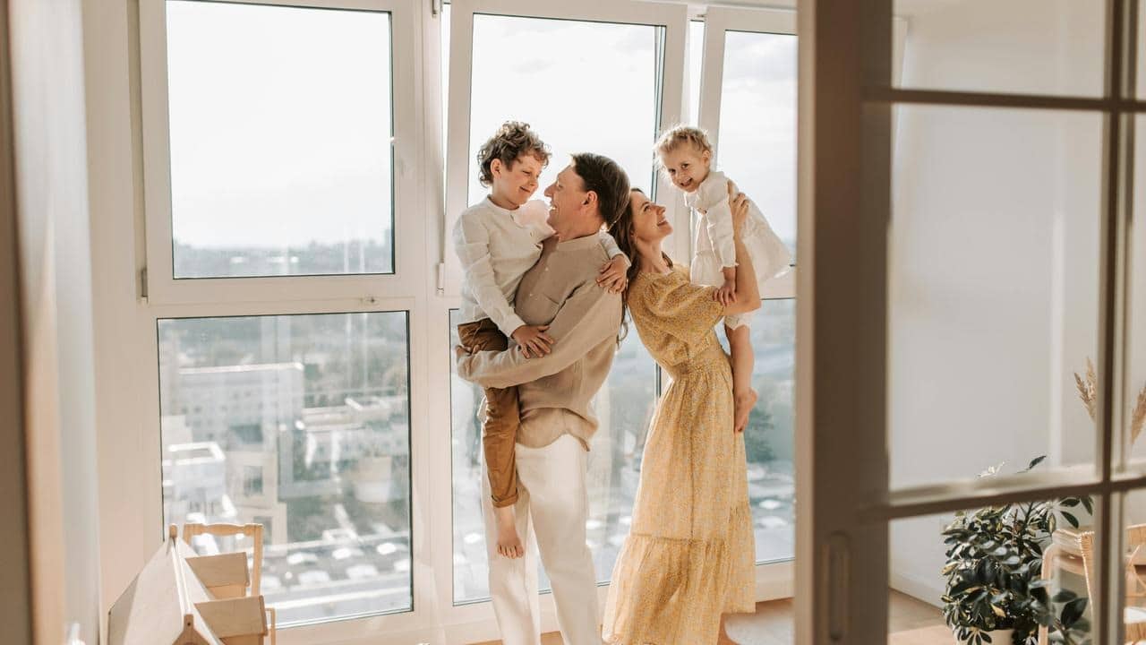 A family smiling and holding two children in a bright room with large windows.
