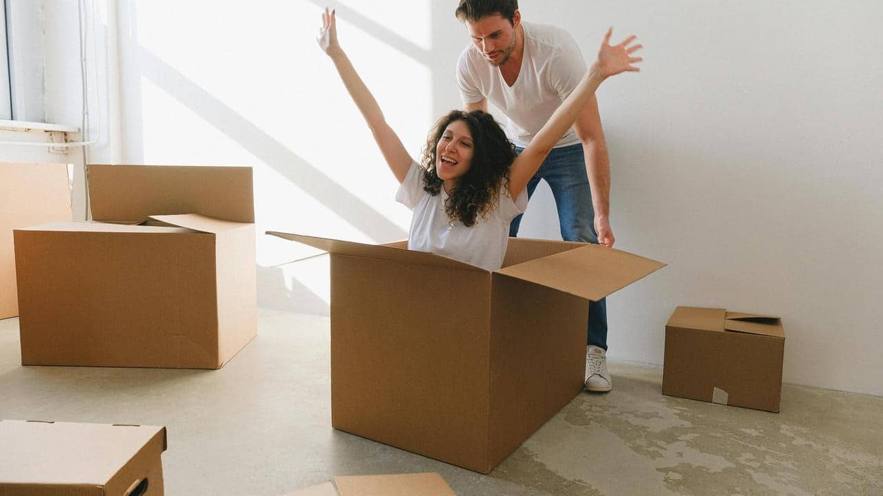 A woman joyfully sitting in a cardboard box as a man stands behind her.