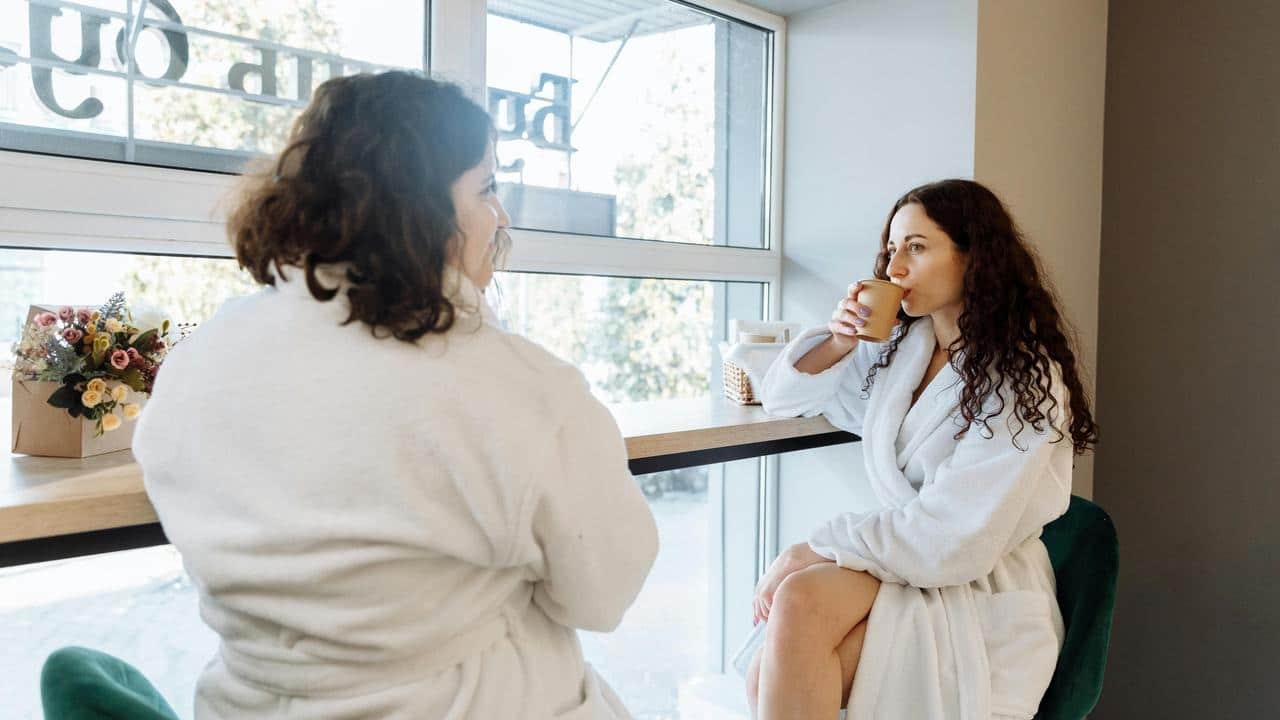 A woman in a robe sipping coffee while talking with another woman by a window.