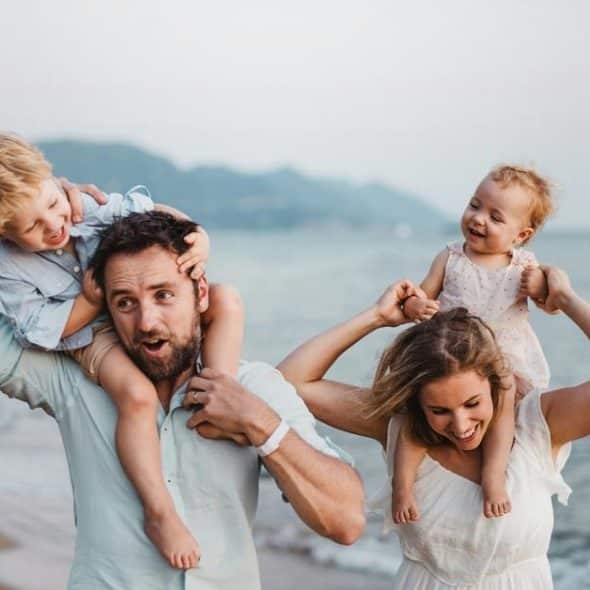 A smiling family of four on a beach, parents carrying young children.