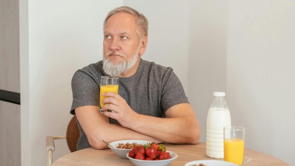 An older man with a white beard sits at a table with juice and fruit.