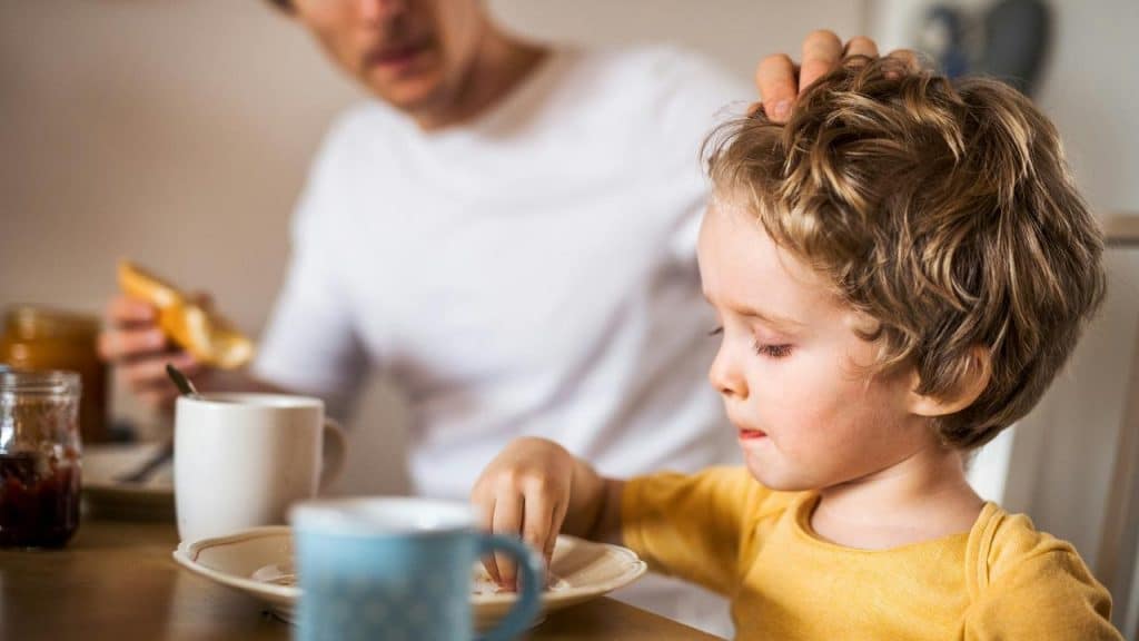 A young child eating breakfast at a table while an adult sits nearby.