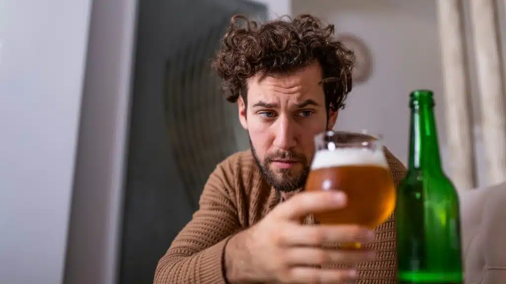 A man with curly hair looks closely at a glass of beer in his hand.