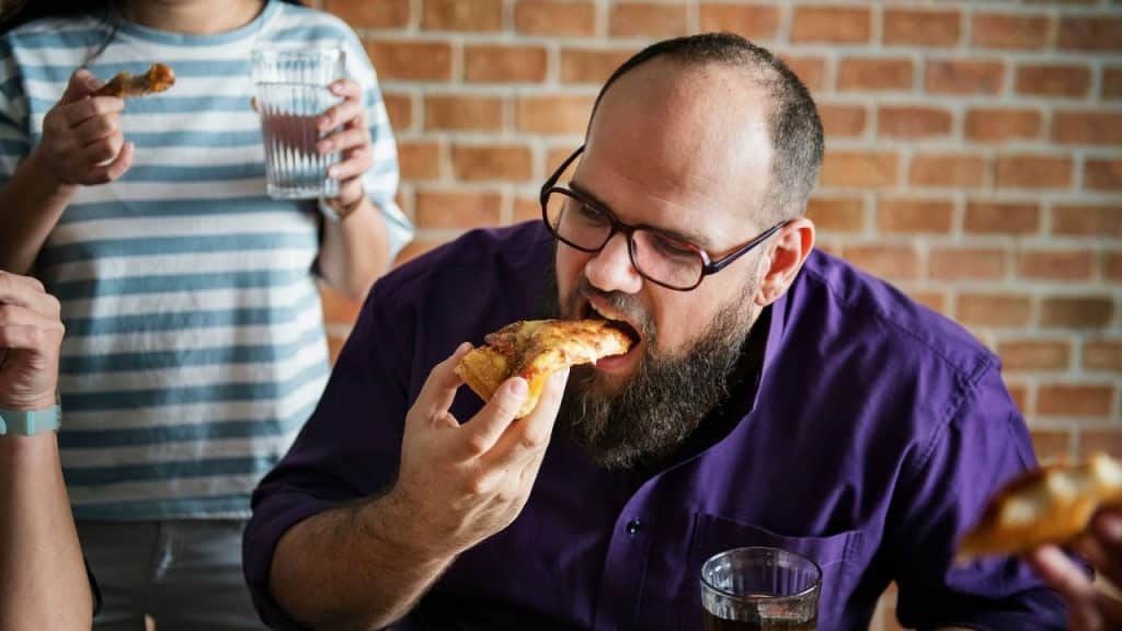 A man with a beard and glasses eats a slice of pizza in a room.