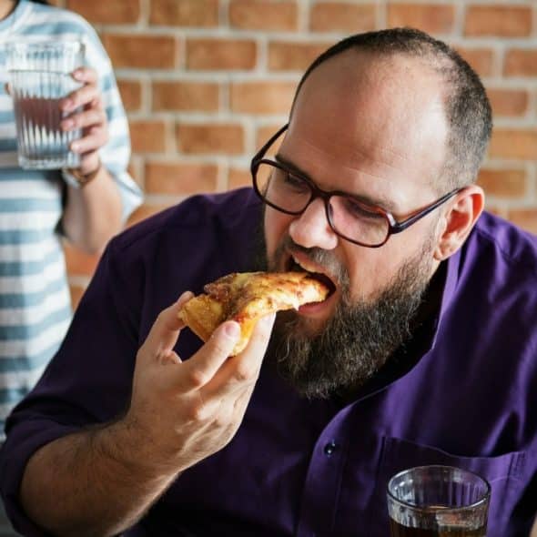A man with a beard and glasses eats a slice of pizza in a room.