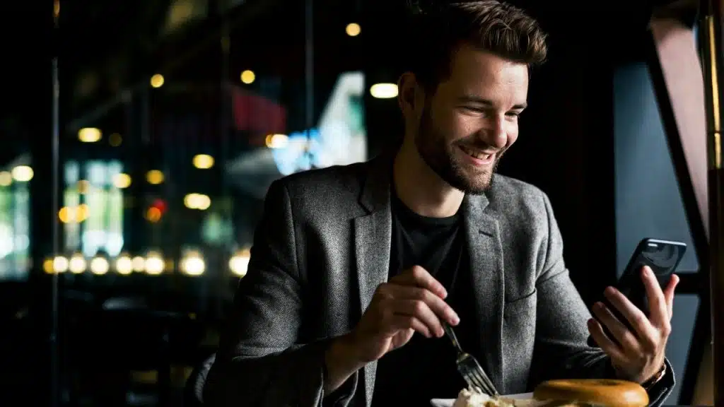 A smiling man in a grey blazer uses a smartphone while sitting at a table.