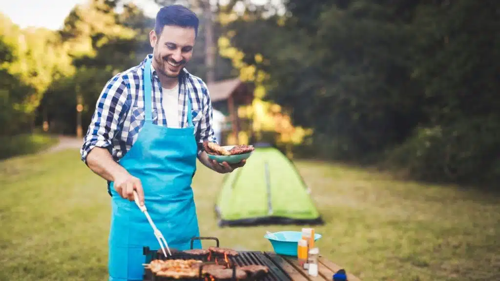 A smiling man in a blue apron and plaid shirt grills meat at a campsite.