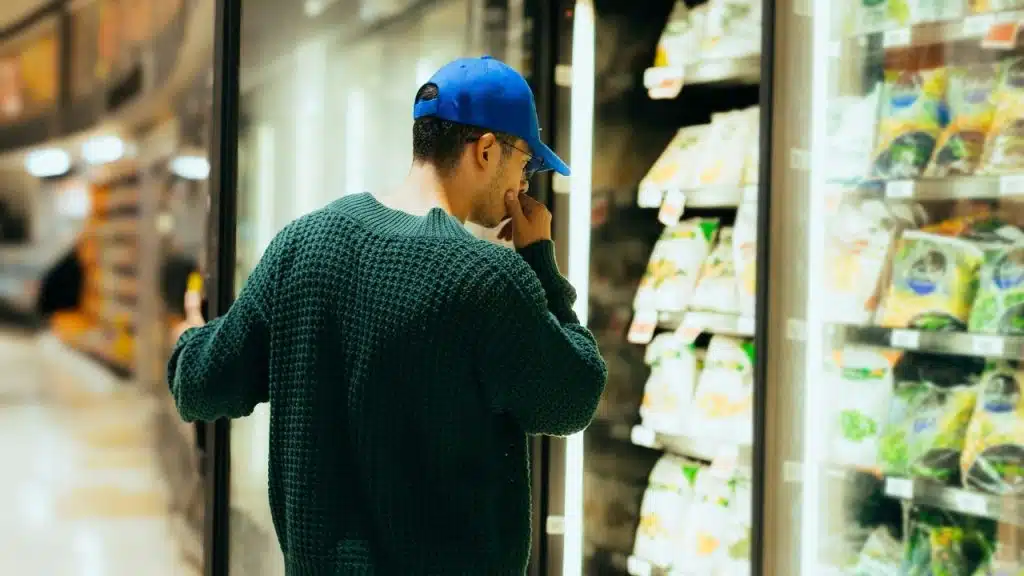 A man wearing a green sweater and blue hat looks at items in grocery refrigerators.
