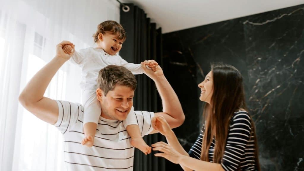 A happy toddler sitting on an adultโs shoulders while another adult smiles nearby indoors.