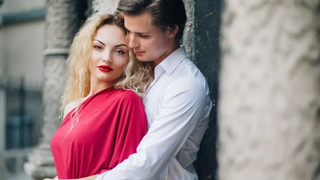 A man in a white shirt embraces a blonde woman wearing a bright red dress.